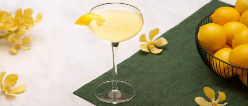A glass of Bee's Knees garnished with lemon peelon a green cloth alog with the yellow flowers  and bowl of lemons 