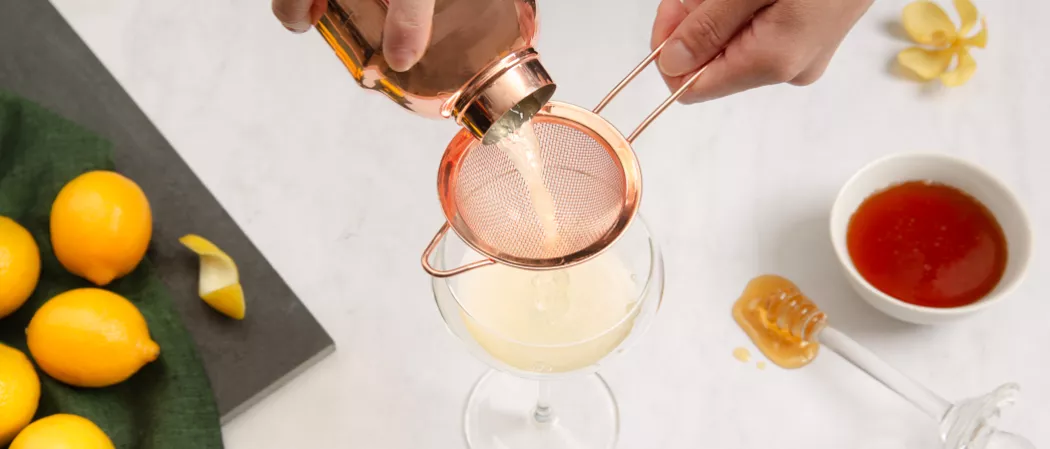 A person holding a fine strainerand pouring lemon juice into a glass along with a  honeydipper, a bowl of honey syrup and some lemons  on a green cloth
