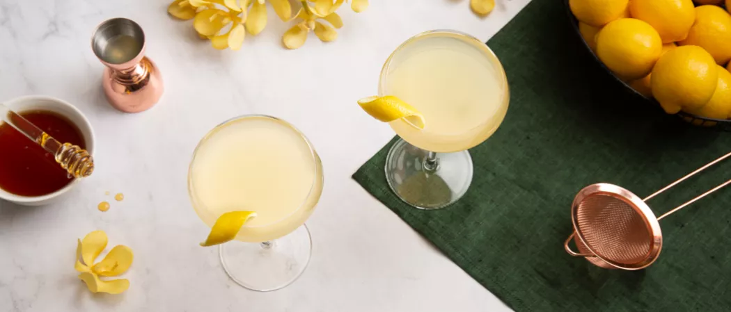 A person holding a strainer and pouring lemon juice into a glass along with a  honeydipper, a bowl of honey syrup and some lemons  on a green cloth