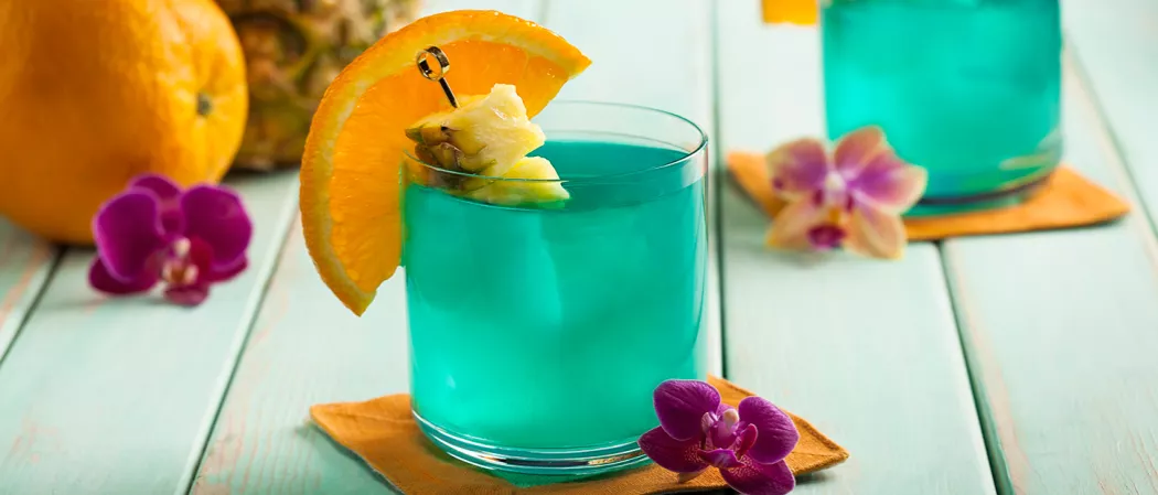 Glass of Blue Hawaii garnished with an orange wedge and a pineapple in pick on a napkin alongside a pink orchid and an orange.