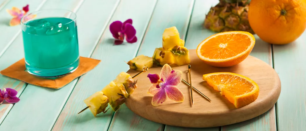 Pineapple slices, pineapples on picks, pink orchids, and some pink flowers on a round wooden chopping board, alongside a glass of Blue Hawaii with ice on an orange napkin and some pink orchids.