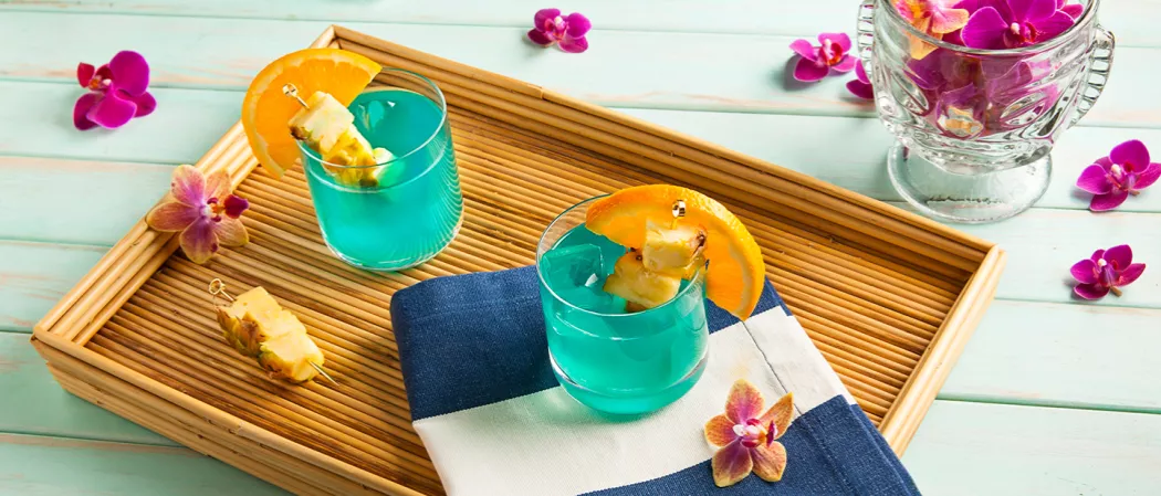 Two glasses of Blue Hawaii garnished with an orange wedge and a pineapple on a pick, one on a napkin  on a wooden tray alongside pink orchids, pineapples in picks, and a bowl with pink orchids.