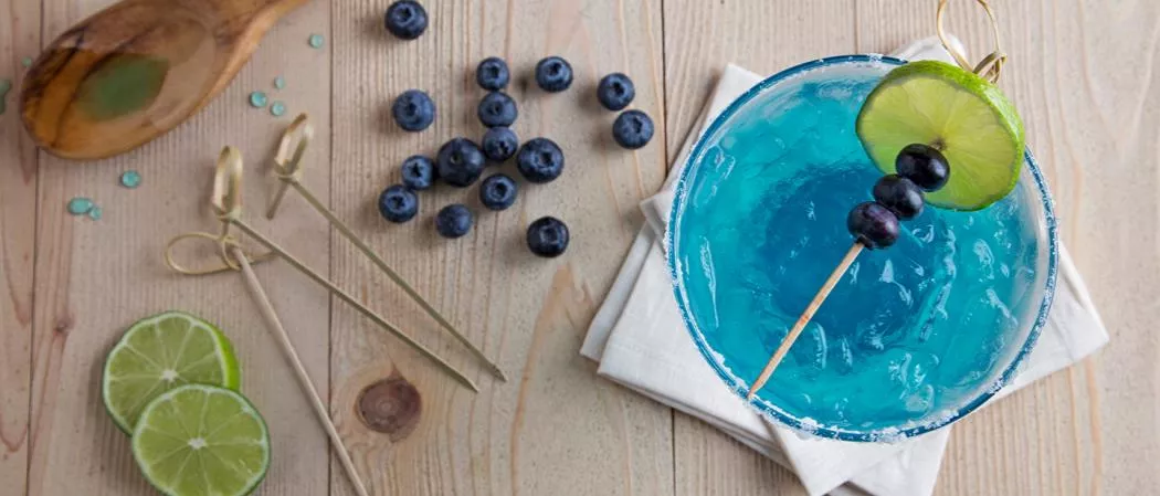 A glass of  Blue Margarita garnished with Lime, Blueberries and Salt along with chopped Lime and Blueberries on table 