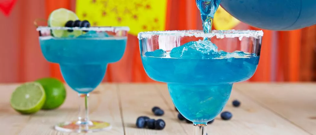 A person Pouring Blue Margarita into a glass and one glass of Blue Margarita is there on the table along with chopped lime and blueberries