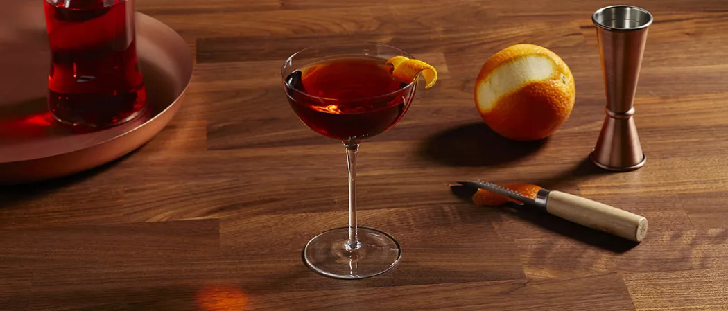 A glass of Basil Hayden® Boulevardier garnished with Orange Peel and half peeled orange and peeler knife on a table