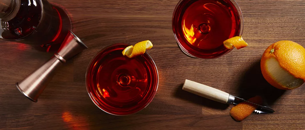 Two glasses of Basil Hayden® Boulevardier garnished with Orange Peel and half peeled orange and peeler knife on a table and a cocktail shaker