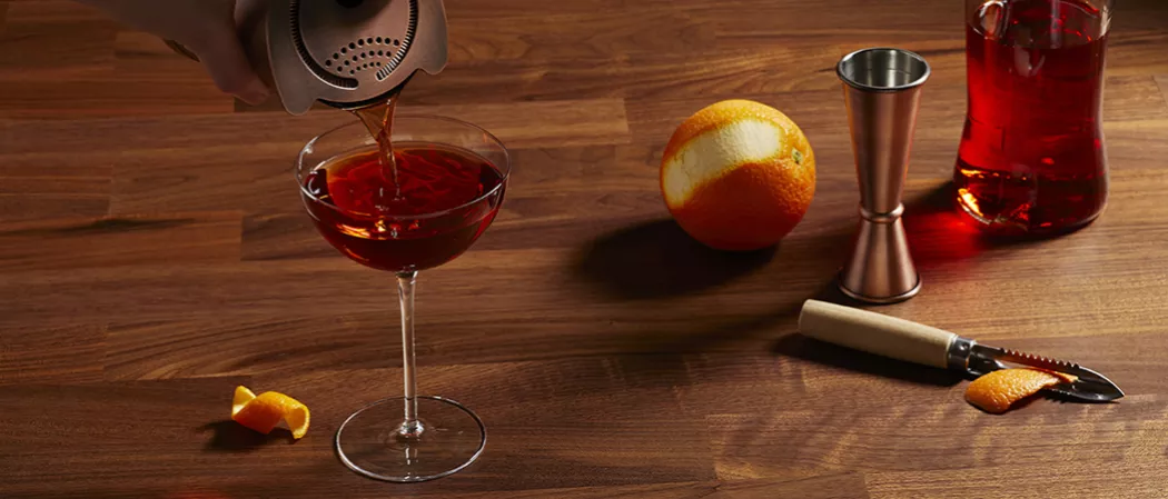 A person pouring Vermouth and Sweet with the help of howthrorne along with half peeled orange and peeler knife on a table and a cocktail shaker