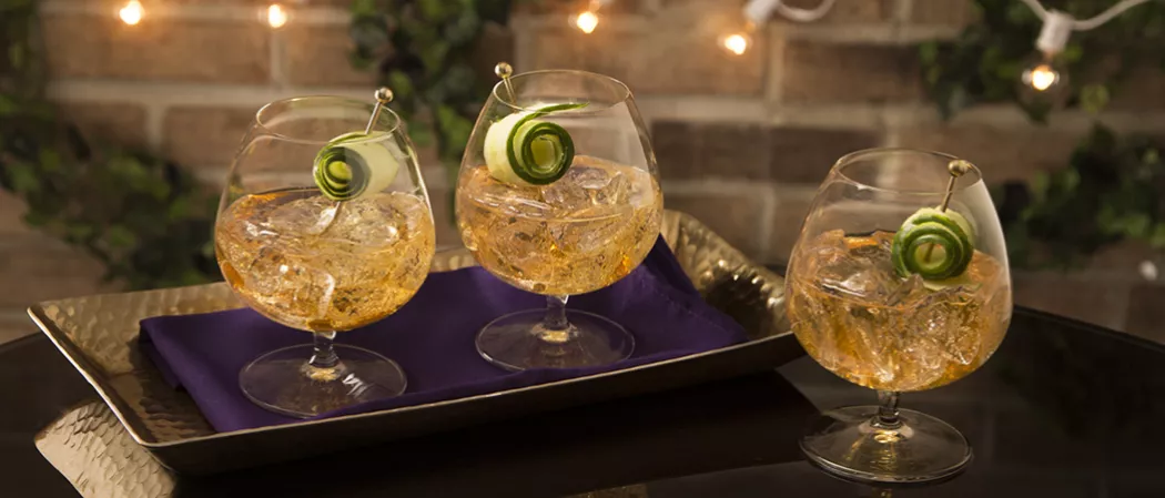Three glasses of Cognac Summit garnished with cucumber peel and crushed ice. Two glasses are on napkins on a tray, and one glass is on the table