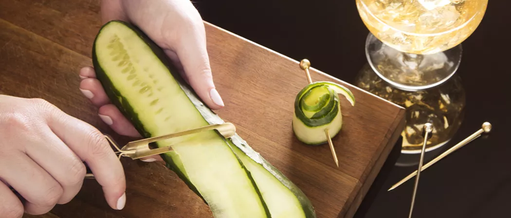 A person using a peeler to peel a cucumber, with cucumber peel nearby