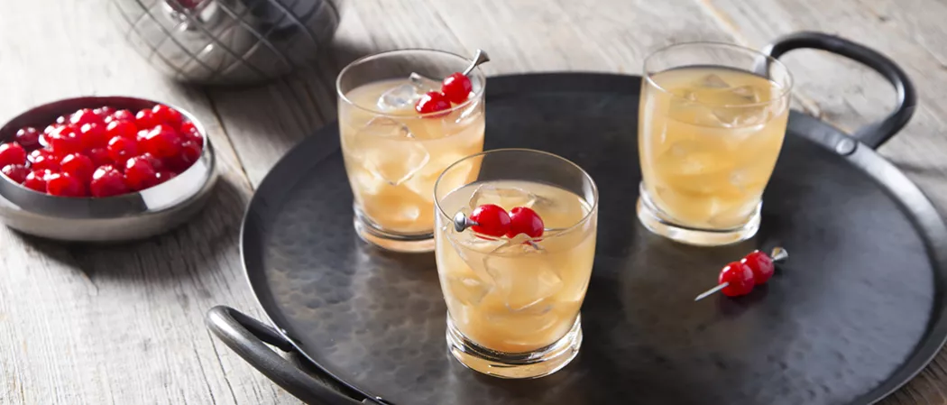 Three glasses of Tropical Spice garnished with cherries on iron tray alongside bowl of cherries