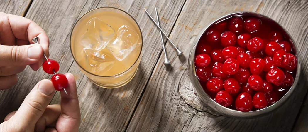 Person putting cherries in steel toothpick alongside glass of Tropical Spice and bowl of cherries