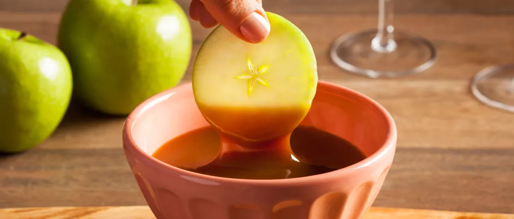A person is dipping an apple wheel into a bowl of DeKuyper® Buttershots® Schnapps, alongside some additional apples