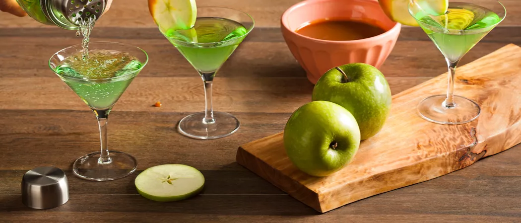 Three glasses of Caramel Apple Martini garnished with an apple wheel.  A  person is pouring Caramel Apple Martini  from a shaker in a glass, on a wooden log with two apples, and the other two are on the table
