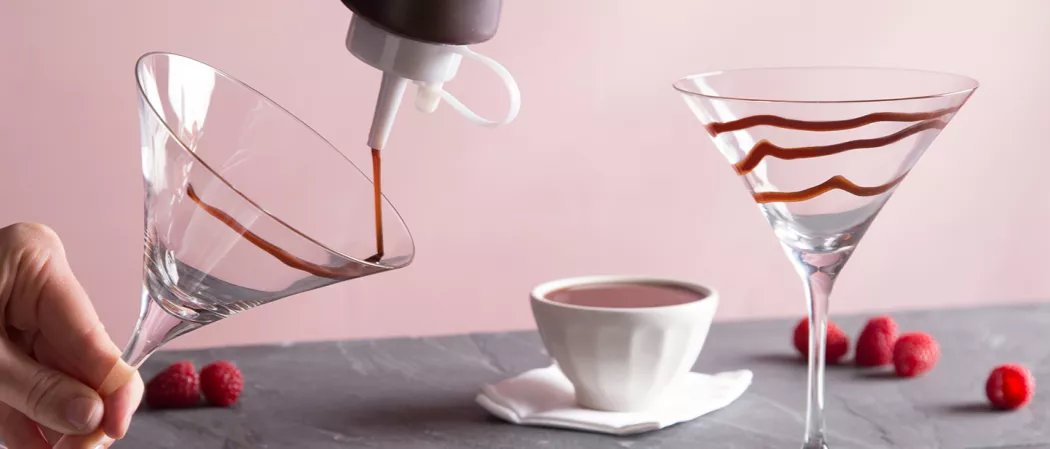 A person is garnishing a glass with chocolate sauce, beside a bowl of chocolate sauce on a white cloth, and another glass garnished with chocolate sauce, alongside a few Raspberries.