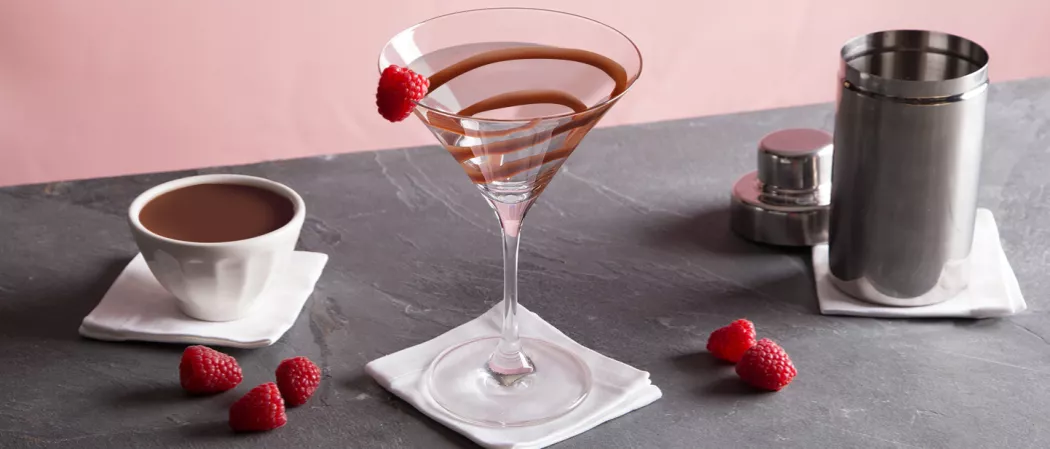 A bowl of chocolate sauce, a glass garnished with chocolate sauce and a Raspberry on rim and a shaker are each arranged on a small piece of white cloth alongside some strawberries.