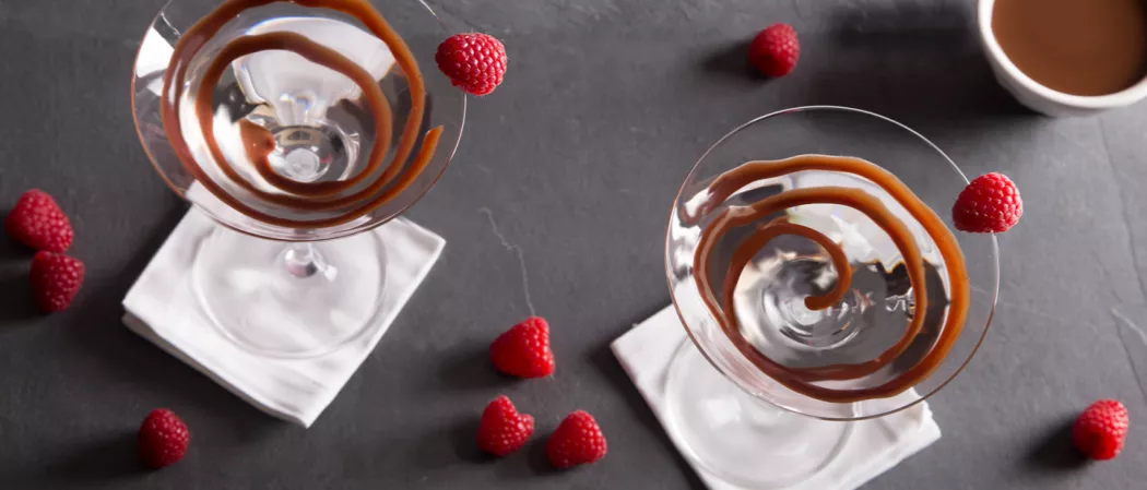 Three glasses, each garnished with chocolate sauce and a Raspberry  on rim are placed on white cloth alongside some Raspberries, and a bowl of chocolate sauce.