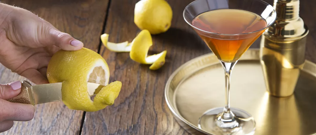 Person peeling lemon alongside Glass of Courvoisier® Sidecar on bronze plate alongside a shaker