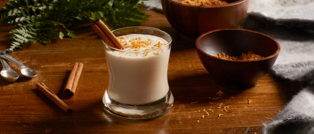 A glass of Cruzan® Coquito garnished with Cinnamon Stick and Shredded Coconut along with shredded coconut bowl  and cinnamon stick on the  table 