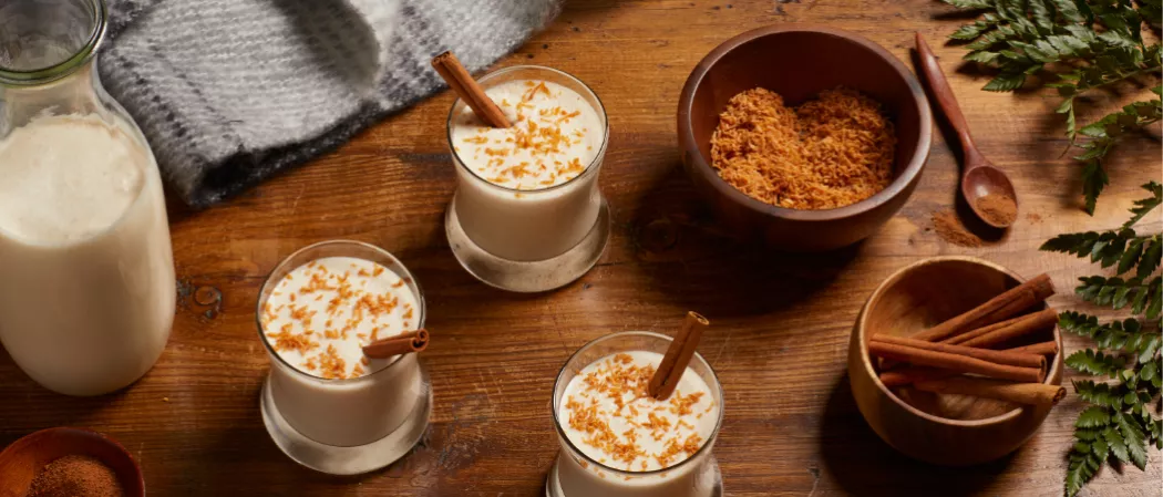 Three glasses of Cruzan® Coquito garnished with Cinnamon Stick and Shredded Coconut along with shredded coconut bowl  and cinnamon stick on the  table  and Sweetened Condensed Milk jar