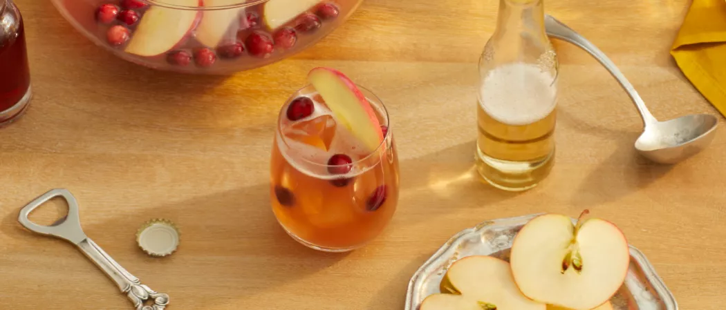 Glass of Cruzan® Cranberry Cider Punch garnished with apple slice and cranberries alongside a plate of apples, bottle opener and ladle