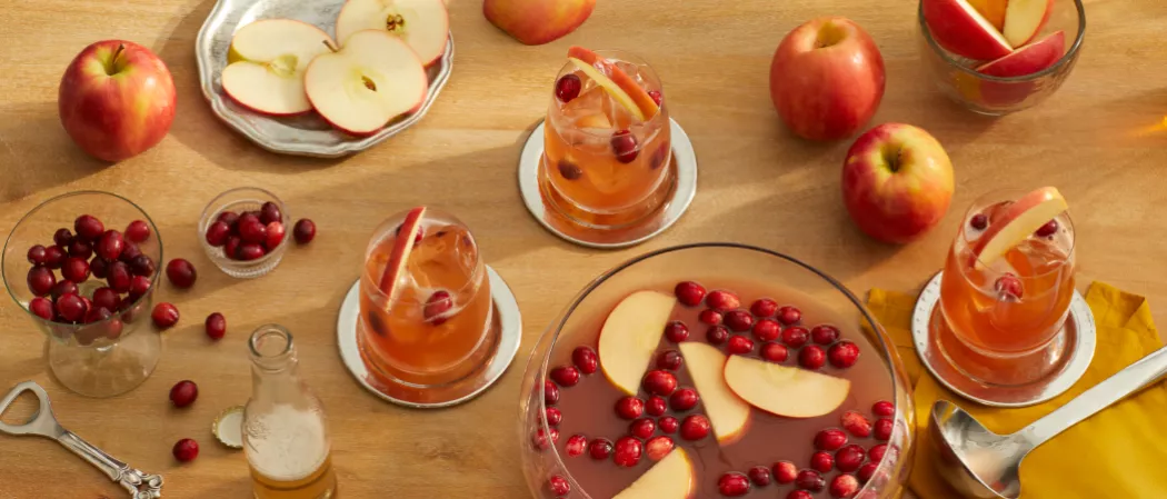 A big bowl and three glasses of Cruzan® Cranberry Cider Punch garnished with apple slices and cranberries on steel coasters,  near by  cranberries, apples, and a bottle opener.