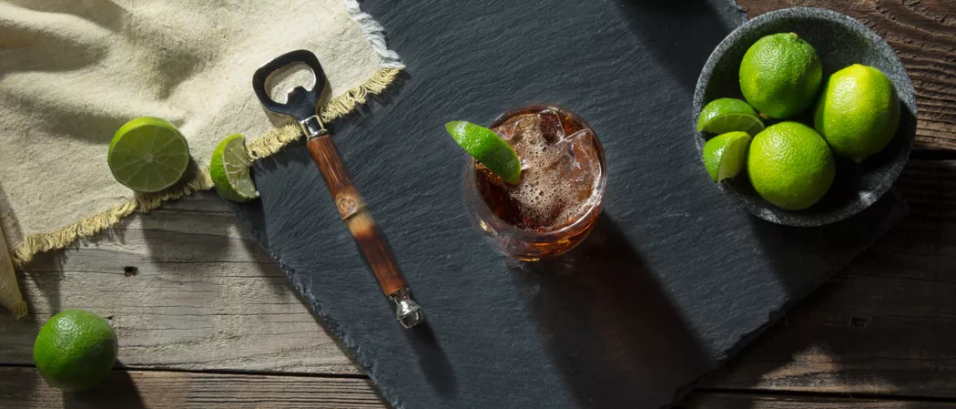 A glass of Cruzan® Safe Harbor garnished with  Lime Wedge along with lime on the table and in a bowl and bottle opener