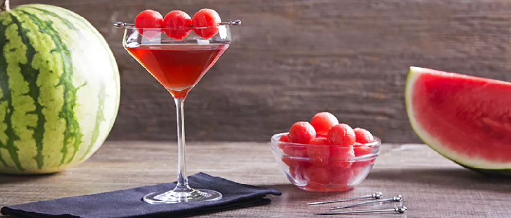 A glass of Melon Creek Martini, garnished with spiked watermelon balls pierced on steel cocktail picks. accompanied by a bowl of spiked watermelon balls and additional steel cocktail picks