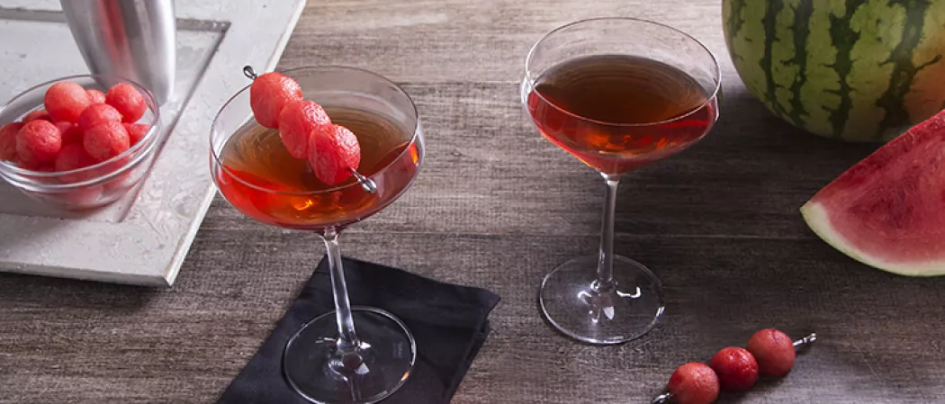 two glasses of Melon Creek Martini, sitting on a cloth, one garnished with spiked watermelon balls pierced on steel cocktail picks, Alongside a bowl full of spiked watermelon balls in a tray