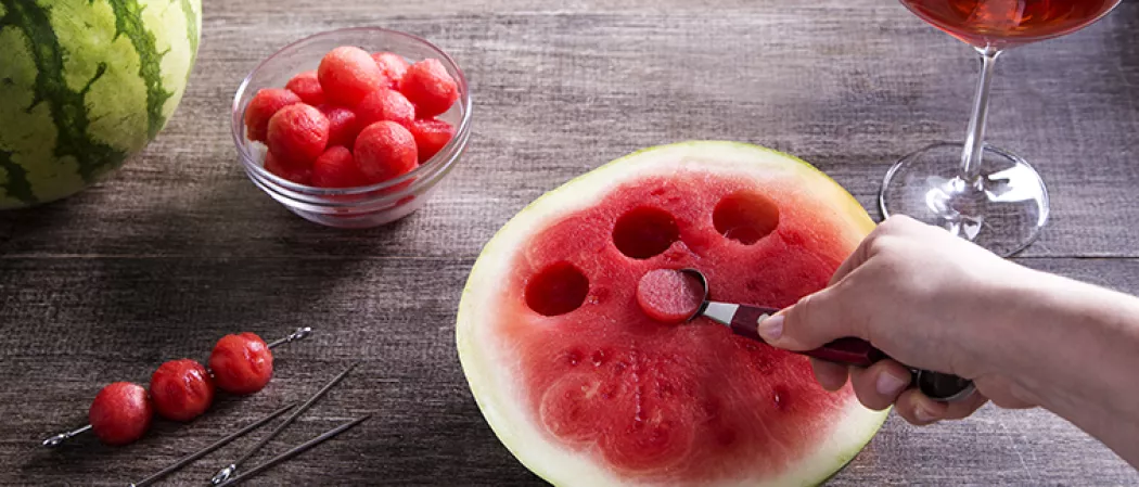 A person is extracting a watermelon ball from the watermelon slice using a portion scoop, alongside spiked watermelon balls on steel cocktail picks, few additional steel cocktail picks and a bowl full of spiked watermelon balls