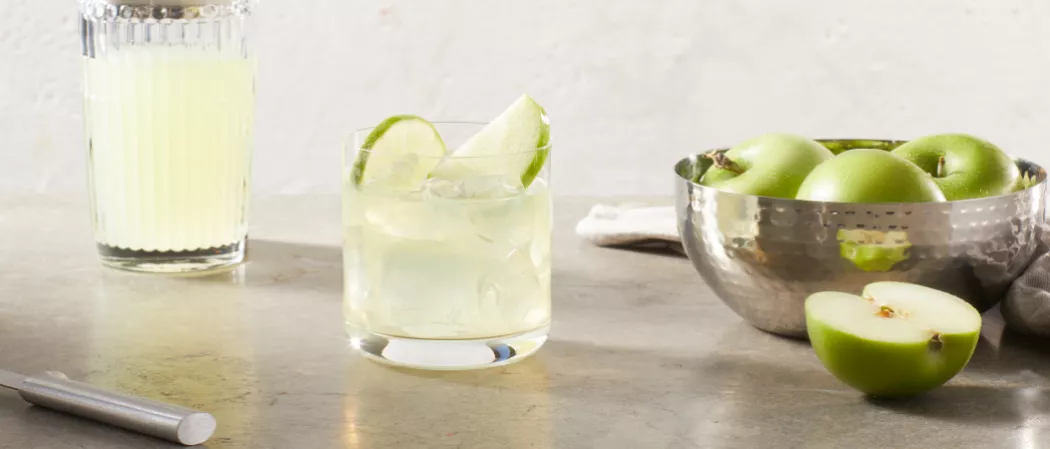 A glass of DeKuyper® End of the Rainbow garnished with Green Apple Slices and Lime Wheel along with bowl of green apple
