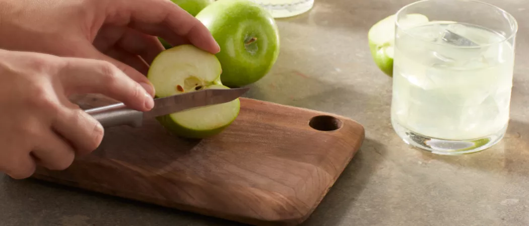A person chopping green apple on a chopping board along with the glass of DeKuyper® End of the Rainbow