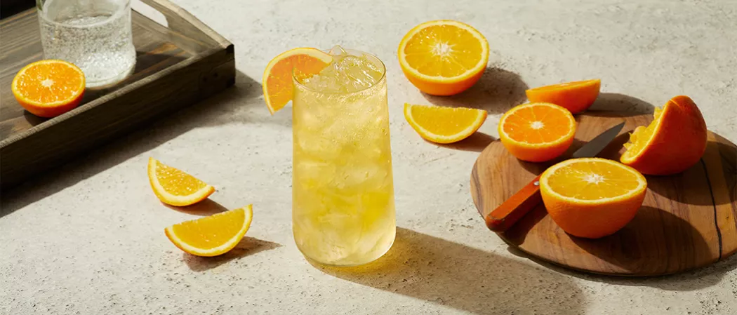 Glass of EFFEN® Blood Orange Crush with crushed ice garnished with orange slice alongside few orange slices and round wooden board with orange and knife on it.