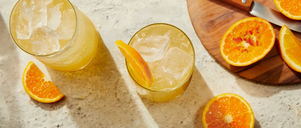 Two glasses of EFFEN® Blood Orange Crush with ice one garnished with orange slice alongside few orange slices and a round wooden board  with orange and knife on it.