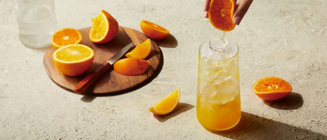 a person squizzing orange juice into glass of EFFEN® Blood Orange Crush alongside few orange slices and round wooden board with orange and knife on it.