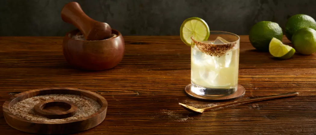 A glass of El Tesoro® Tommy's Margarita garnished with Lime,Spicy Salt along with w wooden plate containg spicy salt and a bowl of chopped lemons 