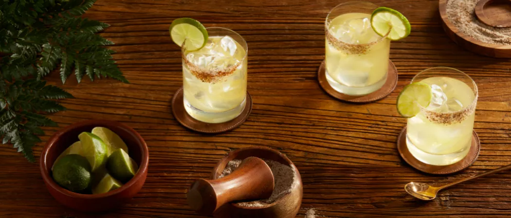 Three glasses of El Tesoro® Tommy's Margarita garnished with Lime,Spicy Salt along with w wooden plate containg spicy salt and a bowl of chopped lemons 