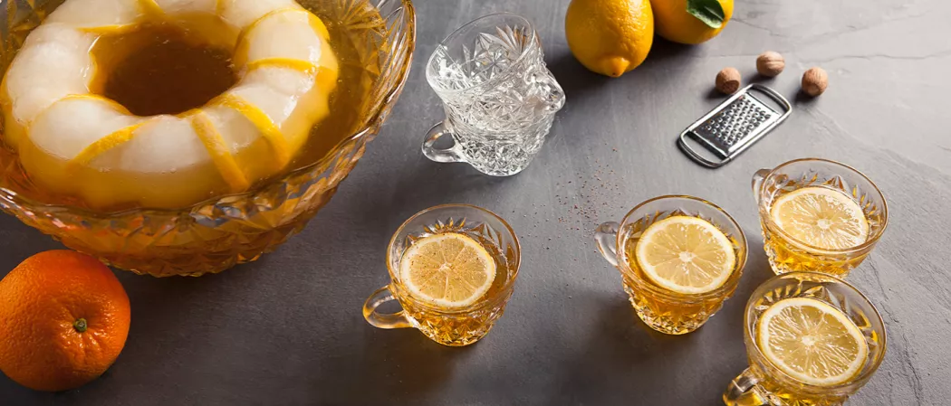 Four cups of Fancy Bourbon Punch garnished with lemon slice alongside a orange, empty cups, grater, some nutmeg and a lemon