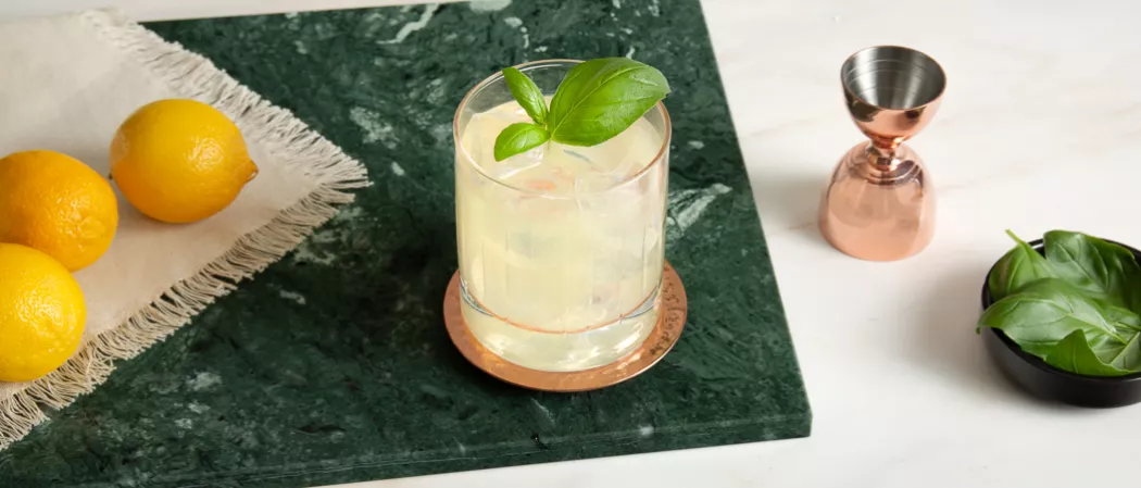 A glass of Gin Basil Smash garnished with mint leaves on a marble counter along with three lemons on a white cloth , a jigger and a bowl of mint leaves