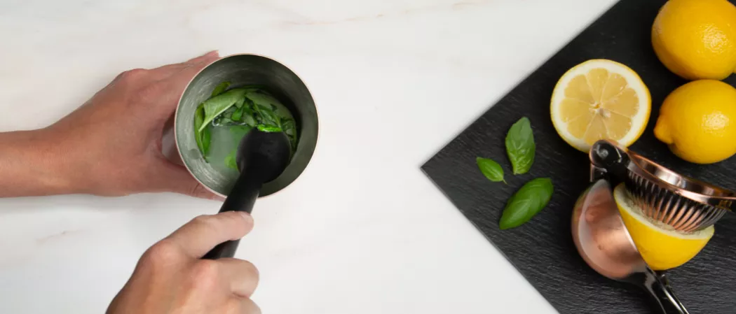 A person holding a muddler using it on lemon juice and mint leaves in a glass  along with the citrus juicer and lemons on a cloth on the kitchen slab