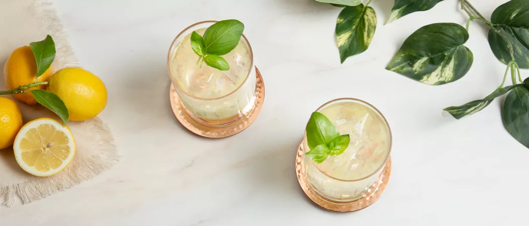 Two glasses of Gin Basil Smash garnished with mint leaves on a copper coaster along with the lemons and mint leaves on a cloth on the kitchen slab