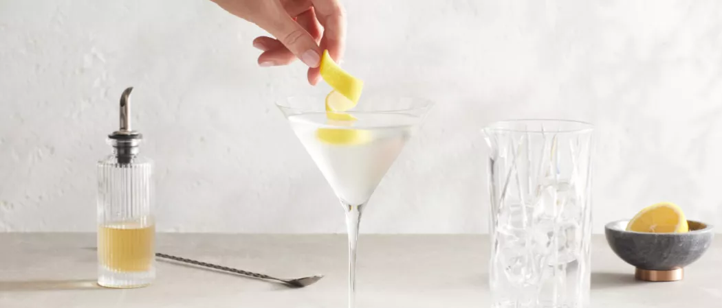 A person garnishing lemon peel on a glass of Haku® Martini along with the bottle of  Orange Bitters