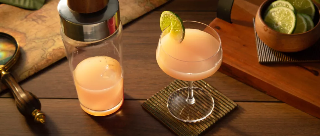 A glass of Hemingway Daiquiri garnished with Lime Wheel and a bottle of Grapefruit Juice