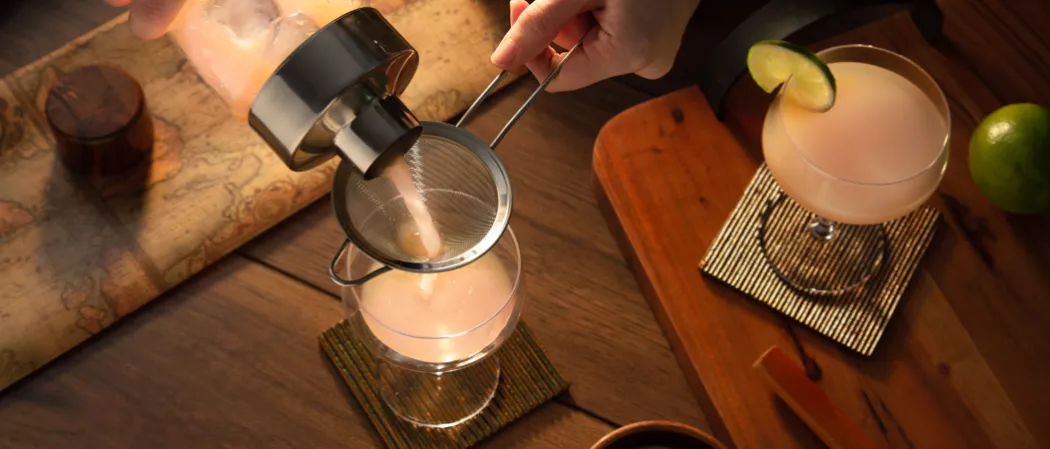 A person pouring grapefruite juice into the glass using fine stariner along with a glass of Hemingway Daiquiri