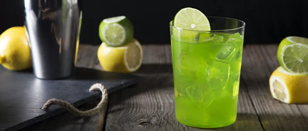 A glass of Hornitos® Midori® Margarita garnished with lemon slice alongside few lemon and limes and a black tray with a glass and lemon on it