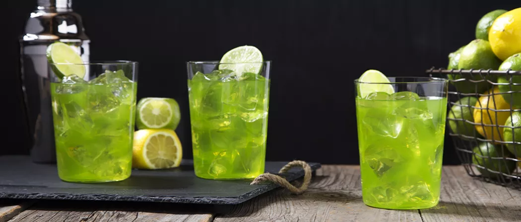 Three glasses of Hornitos® Midori® Margarita garnished with lemon slices two on a black tray alongside basket of lemon and limes.
