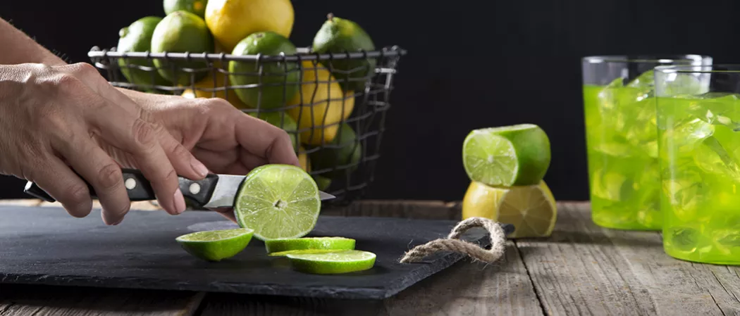 A person cutting slices of lime on a black tray alongside a basket of lemon and limes and two glasses of Hornitos® Midori® Margarita