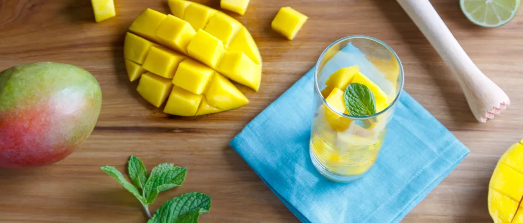 A glass of Mango Mojito garnished with mint and mango slices, alongside fresh mango, mint leaves, chopped mango, and slices.