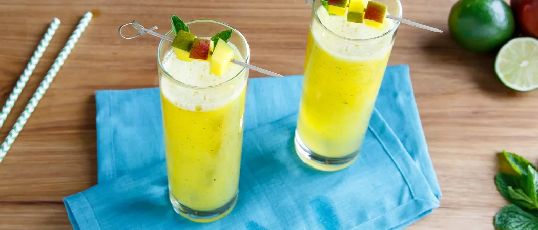 Two glasses of Mango Mojito, each garnished with mint and fruit slices on a cocktail pick, placed on a blue cloth, alongside by fresh mint and straws