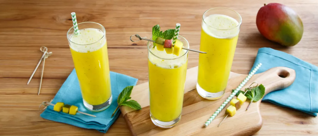 Two glasses of Mango Mojito, one garnished with mint and fruit slices on a pick, alongside fresh mint and fruit slices on picks, placed on a chopping board. Another glass on a blue cloth with fruit slices on picks, accompanied by extra picks.