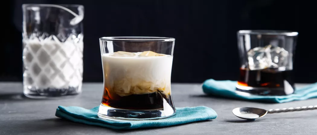 A glass of Coffee Liqueur and cream with ice, and another glass of Coffee Liqueur with ice, both on blue napkins. Alongside a spoon and a jar of cream